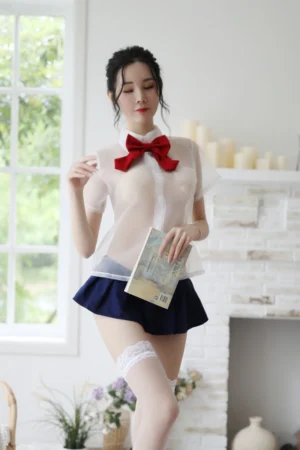 Model posing in b2b student fantasy lingerie featuring a sheer white blouse with red bow and navy pleated skirt