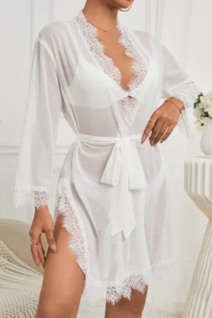Model wearing a white sheer robe style private label sexy see through nightgown with lace edges, long sleeves, and adjustable waist tie.
