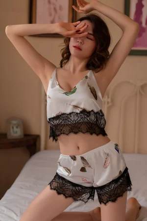 Model wearing a wholesale white lace pajama set featuring a feather print crop top with black lace trim and matching lace shorts in a bedroom setting