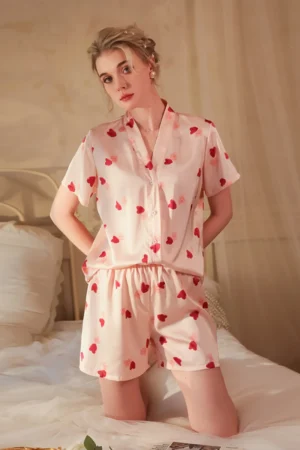 Model wearing a wholesale women's light pink sexy nightgown pajama set with short sleeves, button-front top, and matching heart-print shorts in a bedroom setting.