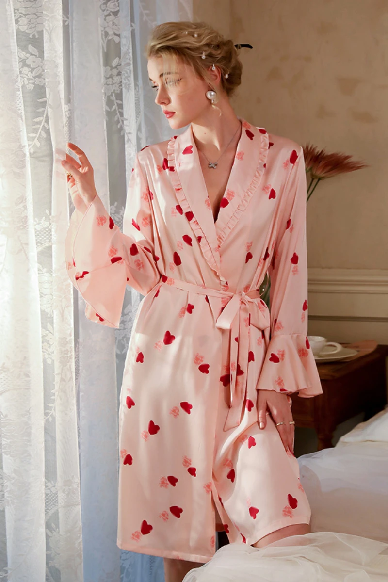 Model wearing a women's light pink sexy nightgown with heart pattern, ruffled neckline, long sleeves, and adjustable waist tie near a window